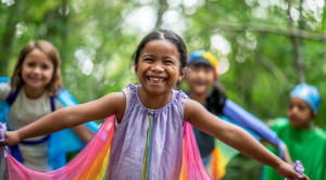 In the forefront, a smiling girl is smiling wearing a cape and pretending to be a dragonfly